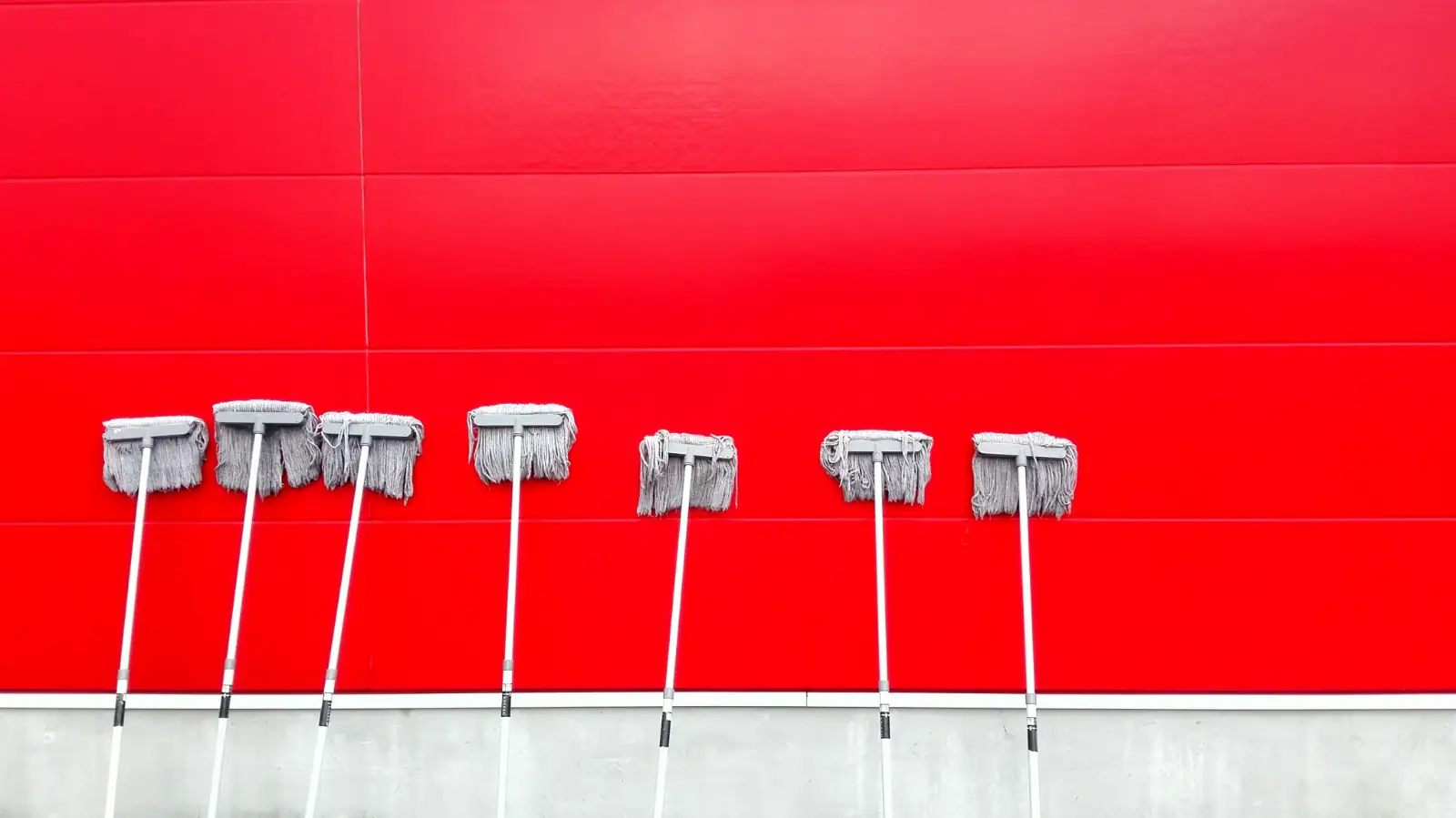 Seven gray mops leaning in a row against a red wall